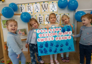 Dzieci trzymają planszę z odbitymi niebieskimi dłońmi.