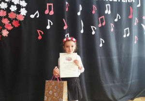 Klara stoi z dyplomem i nagrodą na tle dekoracji konkursowej.