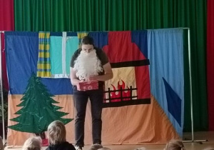 Aktor z brodą Mikołaja otwiera prezent.