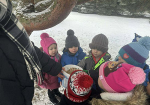 Dzieci zgromadzone w zaśnieżonym ogrodzie stojąc w kole oglądają ziarna dla ptaków