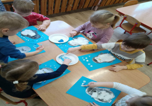 Dzieci siedzą przy stoliku i wykonują pracę plastyczną przedstawiającą pingwina