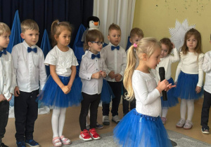 Dziewczynka stoi przed szeregiem dzieci i recytuje wiersz do mikrofonu