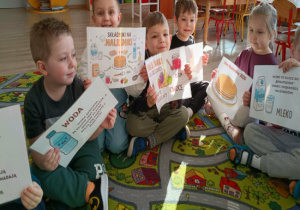 Kilkoro dzieci trzyma plakaty przedstawiające różne składniki potrzebne do zrobienia naleśników