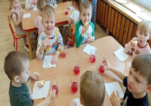 Chłopcy z uśmiechami na twarzach siedzą przy stoliku jedzą pączki i piją sok.