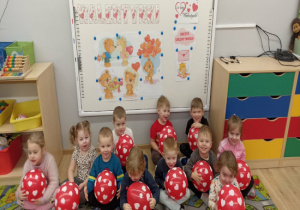 Dzieci siedzą na dywanie na tle napisu walentynki i trzymają czerwone balony w białe serca.