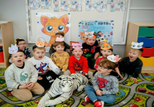 Dzieci siedzą na dywanie w opaskach kotów na głowie i miauczą.