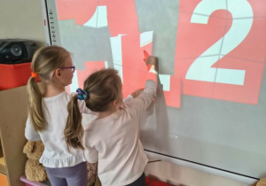 Dzieci układają puzzle na tablicy multimedialnej z numerem alarmowym.