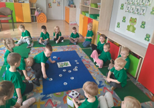 02 Dzieci siedzą w kole na dywanie chłopiec rzucił kostką i karmi papierową żabę odpowiednią ilością przysmaków używając klamerki do prania.