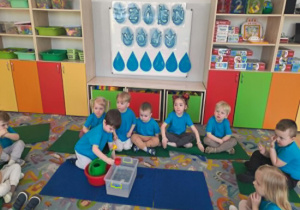 Dzieci siedzą w kole na dywanie chłopiec siedzący na środku przelewa kubkiem wodę z pojemnika do konewki.