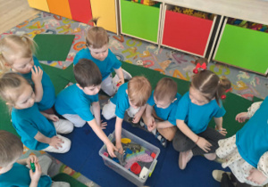 Dzieci siedzą dookoła pojemnika z wodą i wkładają do niego różne przedmioty sprawdzając co zatonie a co będzie pływać.