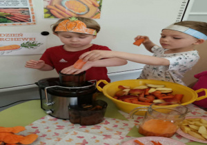 Jeden chłopiec wkłada marchewki do sokowirówki a drugi popycha je dalej specjalnym przyrządem i dzieci patrzą jak sok marchewkowy wypływa do pojemnika.