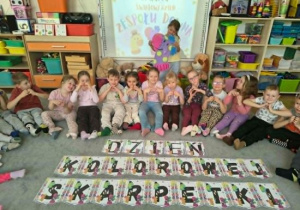 Dzieci siedzą na dywanie z napisem "Dzień Kolorowej Skarpetki"