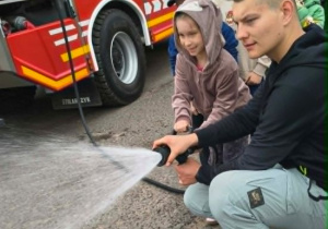 Dzieci wraz z młodym strażakiem biorą udział w pokazie przy wozie strażackim ucząc się obsługi strażackiego węża wodnego.