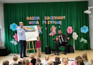Na scenie podczas przedszkolnego koncertu Pan Maciej prowadzi zajęcia dla dzieci a obok akordeonista gra muzykę.